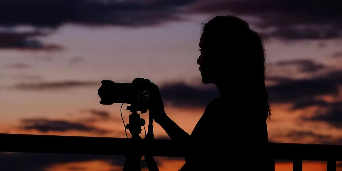 The silhouette of a photographer working at a tripod-mounted camera, rendered as a dark shape against a deep orange and violet dusk sky that fills most of the frame.