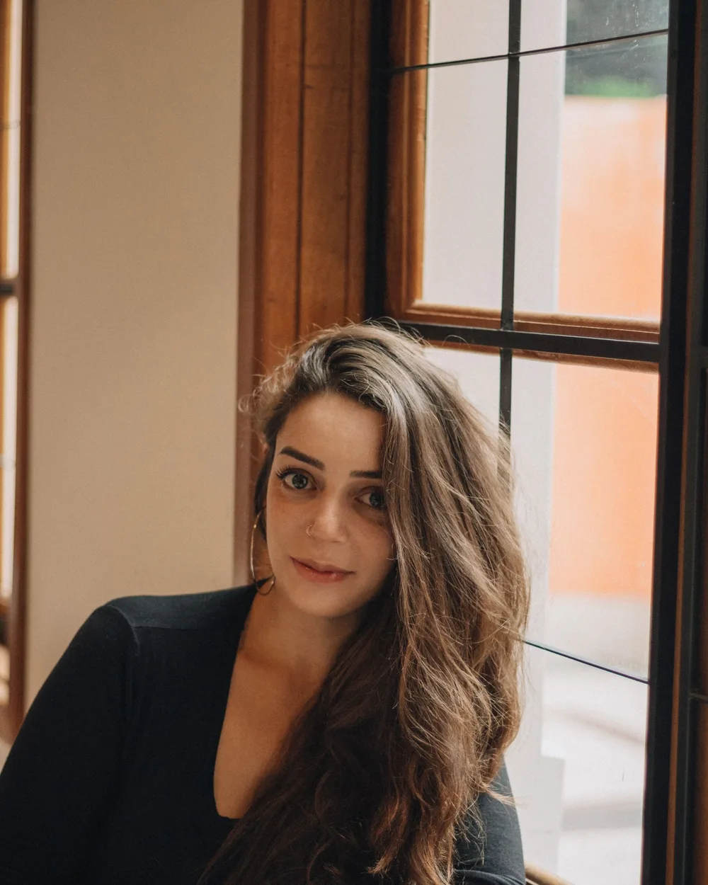 A woman seated beside a tall wood-framed window, her face lit from one side by soft daylight while the far side of her face falls into gentle shadow.